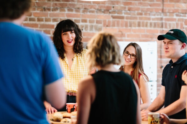 A smiling woman in a yellow shirt standing next to a smiling woman in a red shirt standing around a table with coworkers